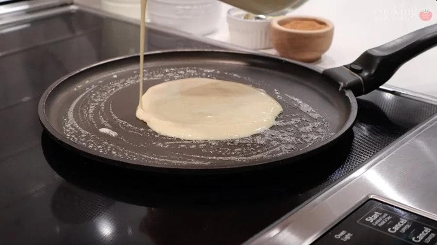 Crepe batter being poured into a buttered non-stick pan on the stovetop.