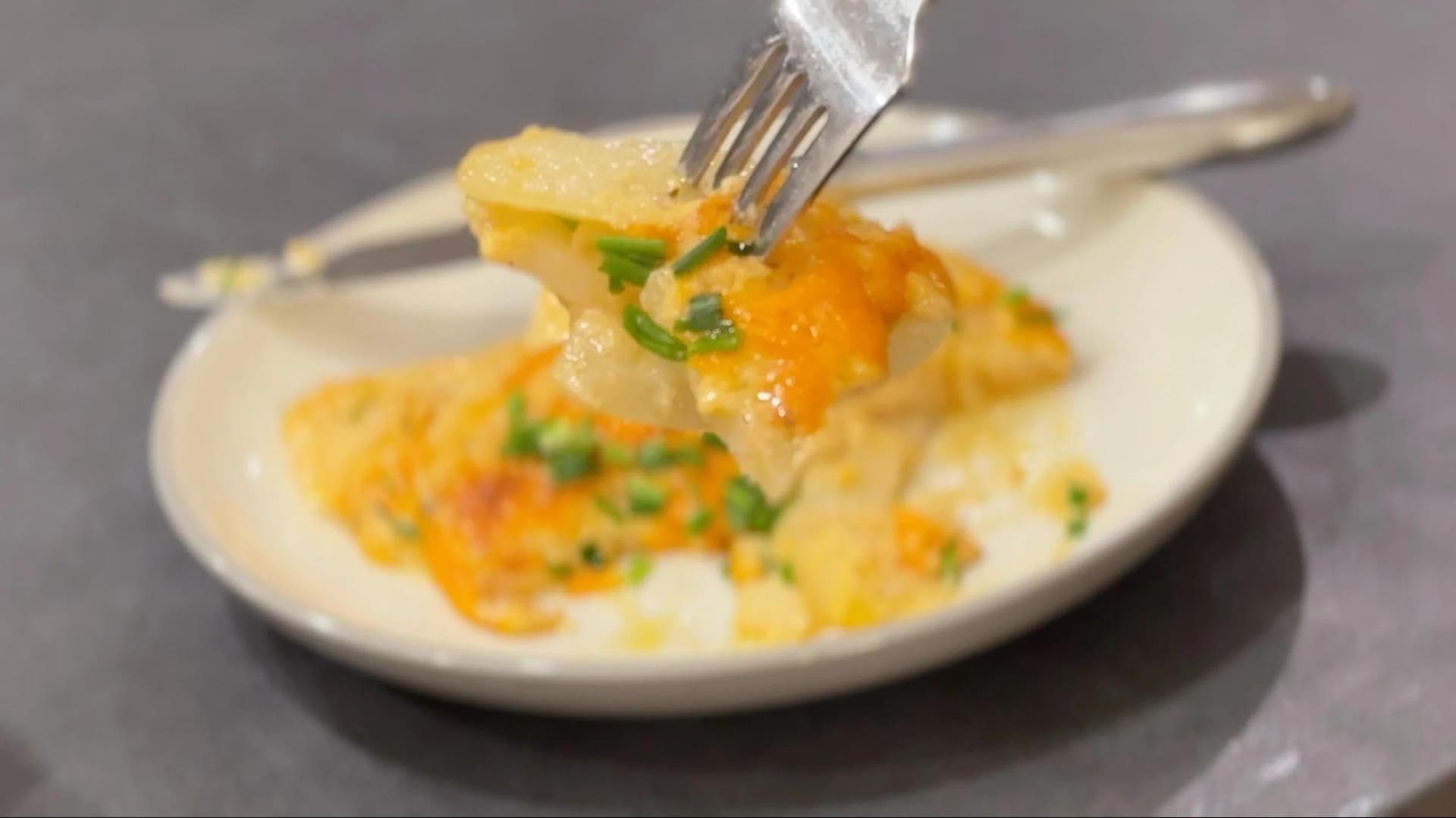 Fork lifting a serving of creamy scalloped potatoes with melted cheese and green garnish from a white plate.