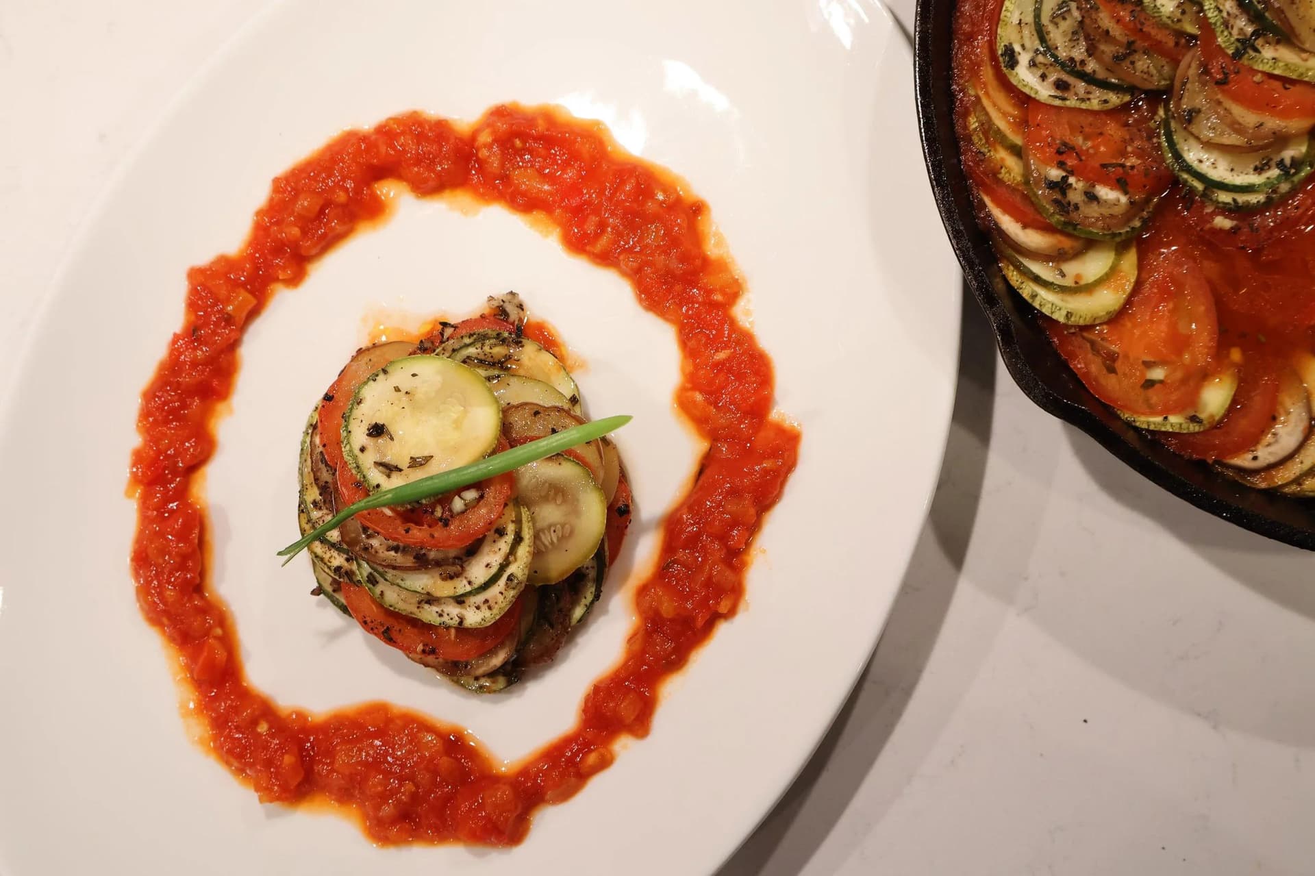 Plated ratatouille stack of layered zucchini, tomato, and eggplant rounds on swirled tomato piperade sauce, garnished with chives.