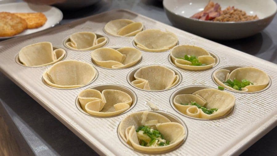 Tortilla cups filled with cheese, vegetables, and fresh herbs arranged on a white plate before baking.