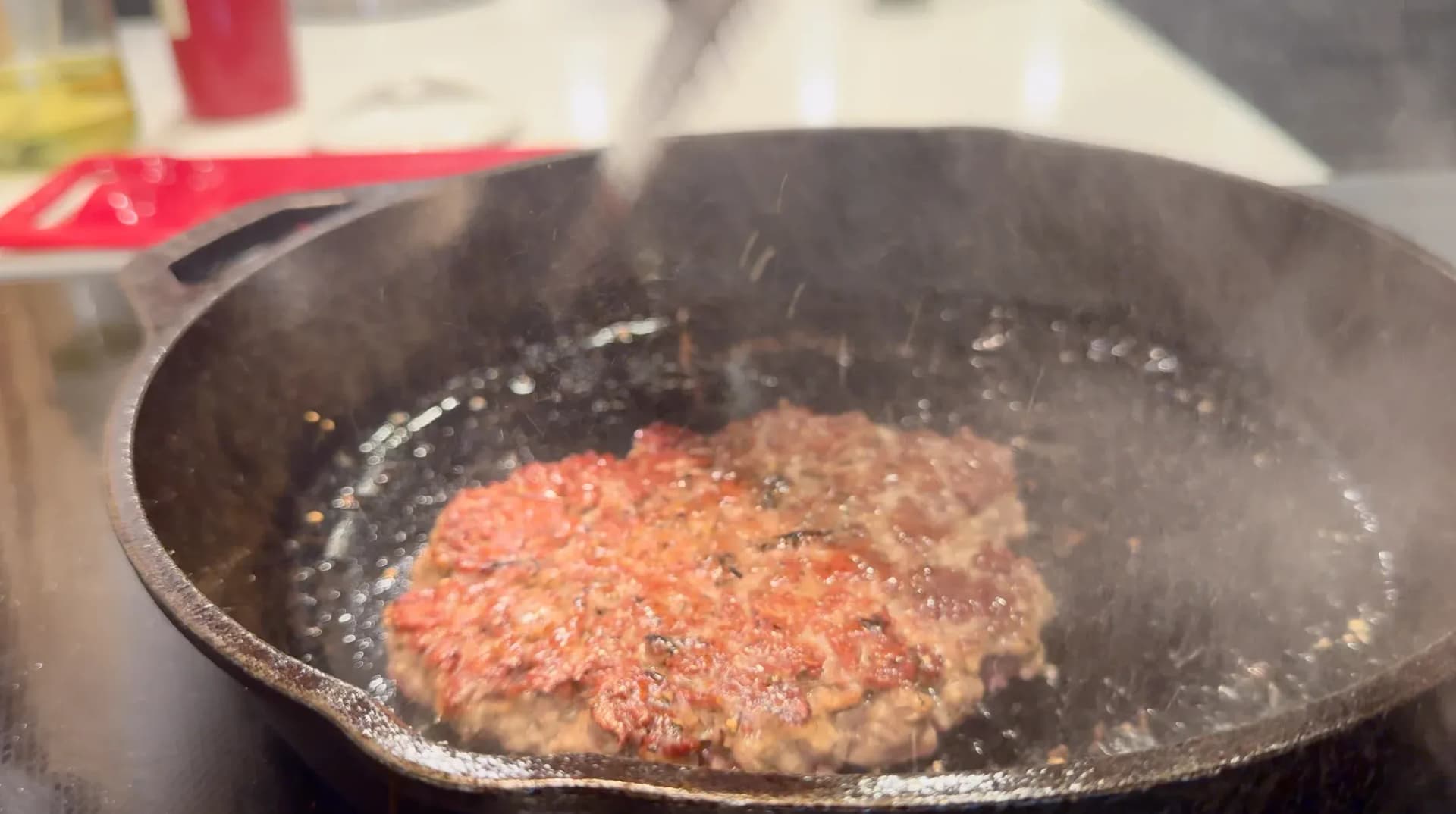 Raw burger patty sizzling in cast iron skillet with oil, seasoned with salt and pepper.