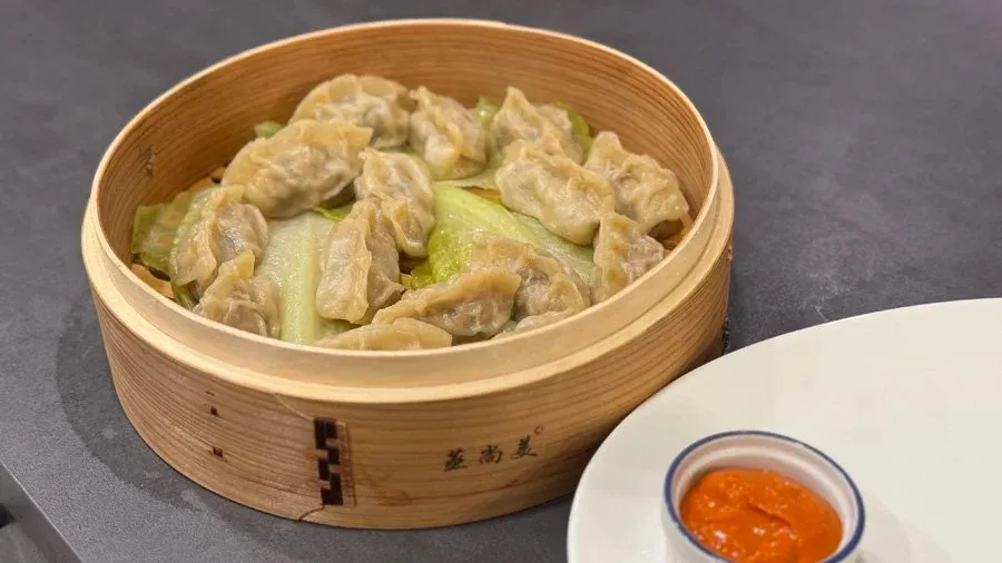 Steamed chicken momos in a bamboo basket with bok choy and a small bowl of orange dipping sauce.