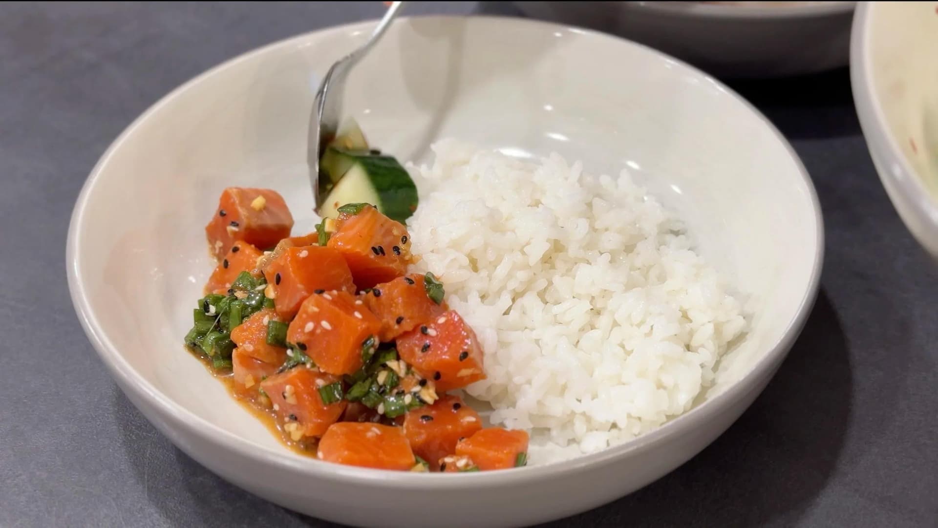 White bowl with fluffy white rice and orange diced salmon poke with green herbs and sesame seeds, fork resting on edge.