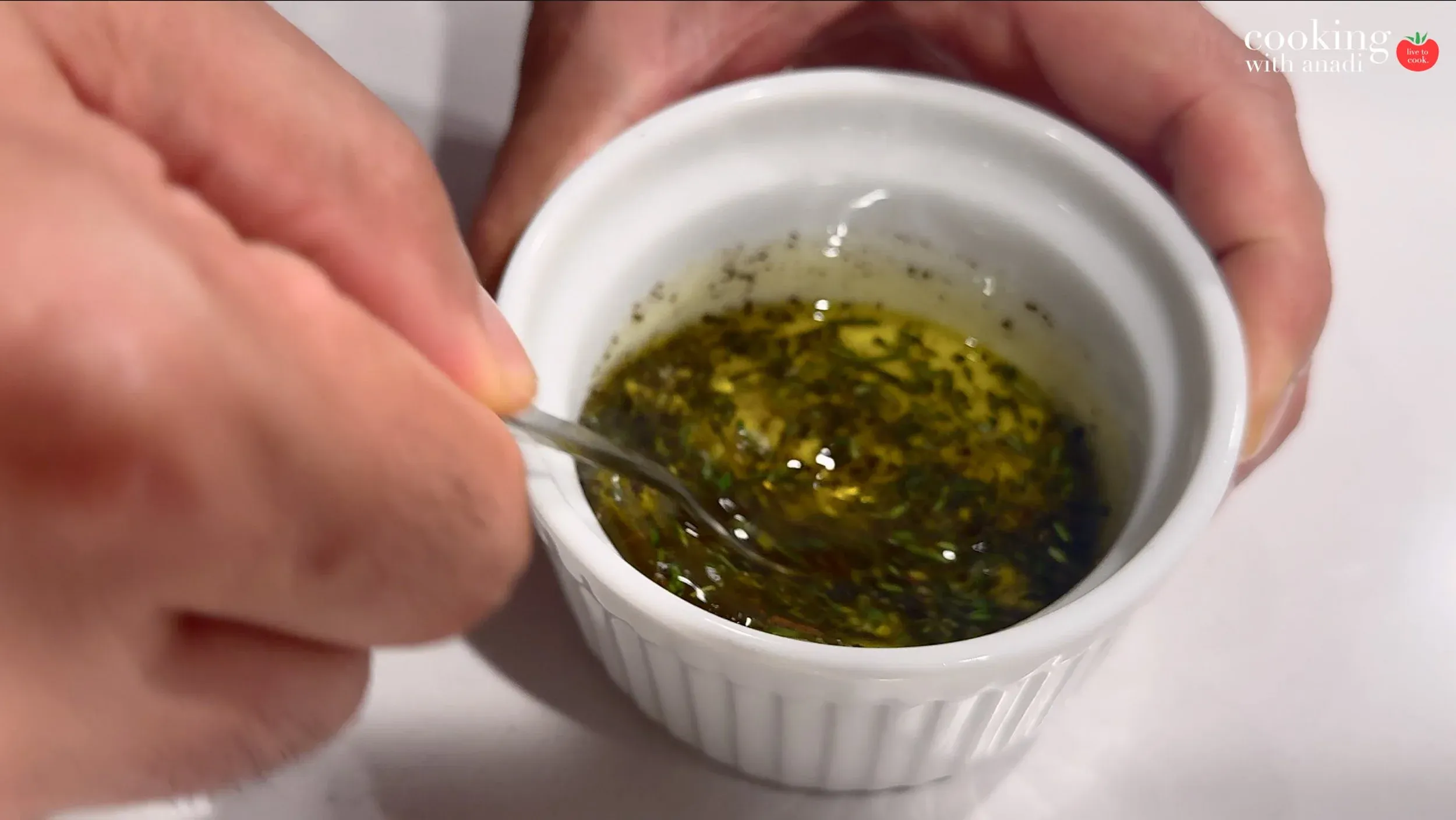 Stirring herb-infused olive oil with garlic in a small white ramekin using a spoon.