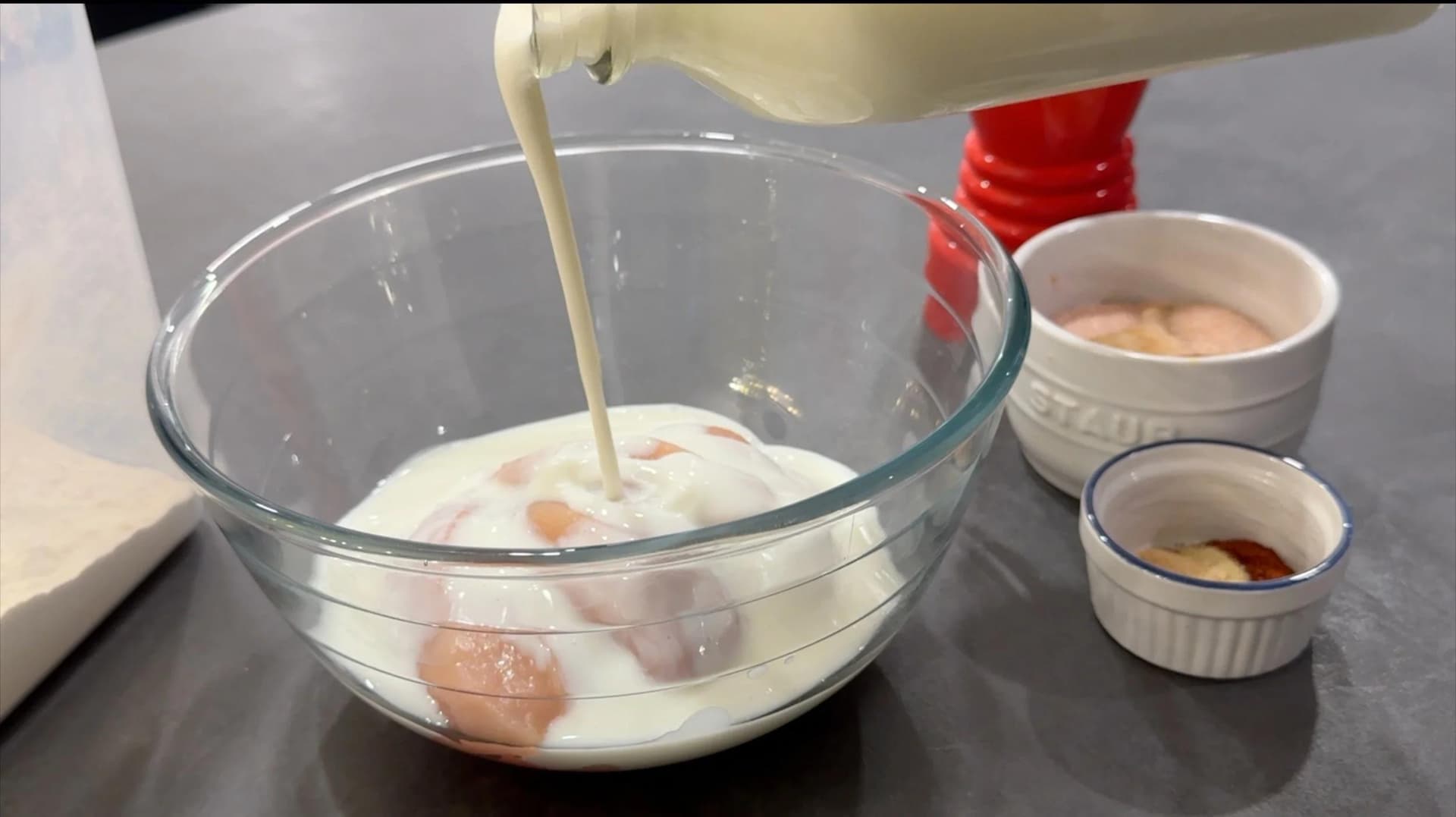 Buttermilk being poured into a glass bowl over spiced chicken tenders for marinating.