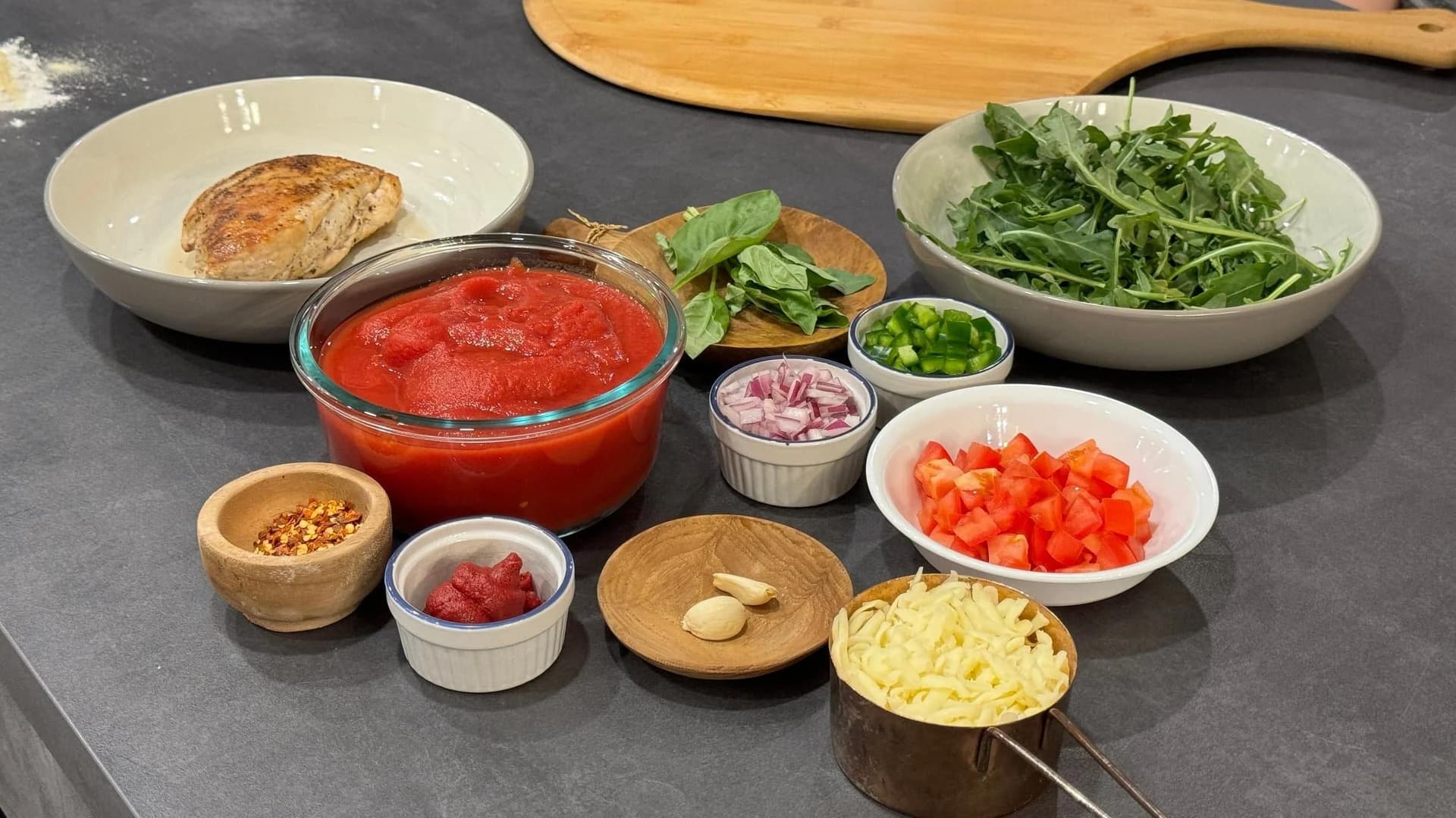 Ingredients laid out for heart-healthy pizza including tomato sauce, fresh arugula, diced tomatoes, garlic, red onion, and cooked chicken breast.