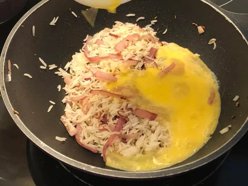 Beaten eggs being poured into a wok with cooked rice and ham pieces, ready to be scrambled together.