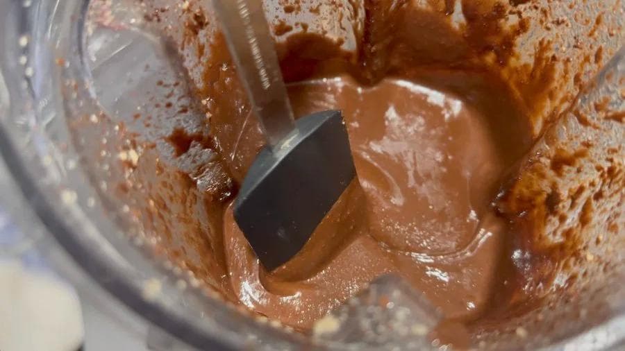 Chocolate hazelnut spread being blended in a food processor with a spatula, showing smooth, creamy texture.