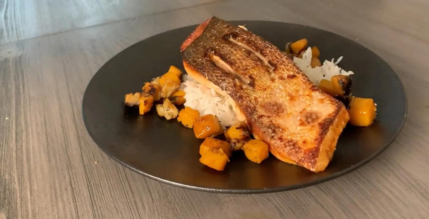 Crispy-skinned salmon fillet served skin-side up on white rice with roasted butternut squash cubes on a dark plate.