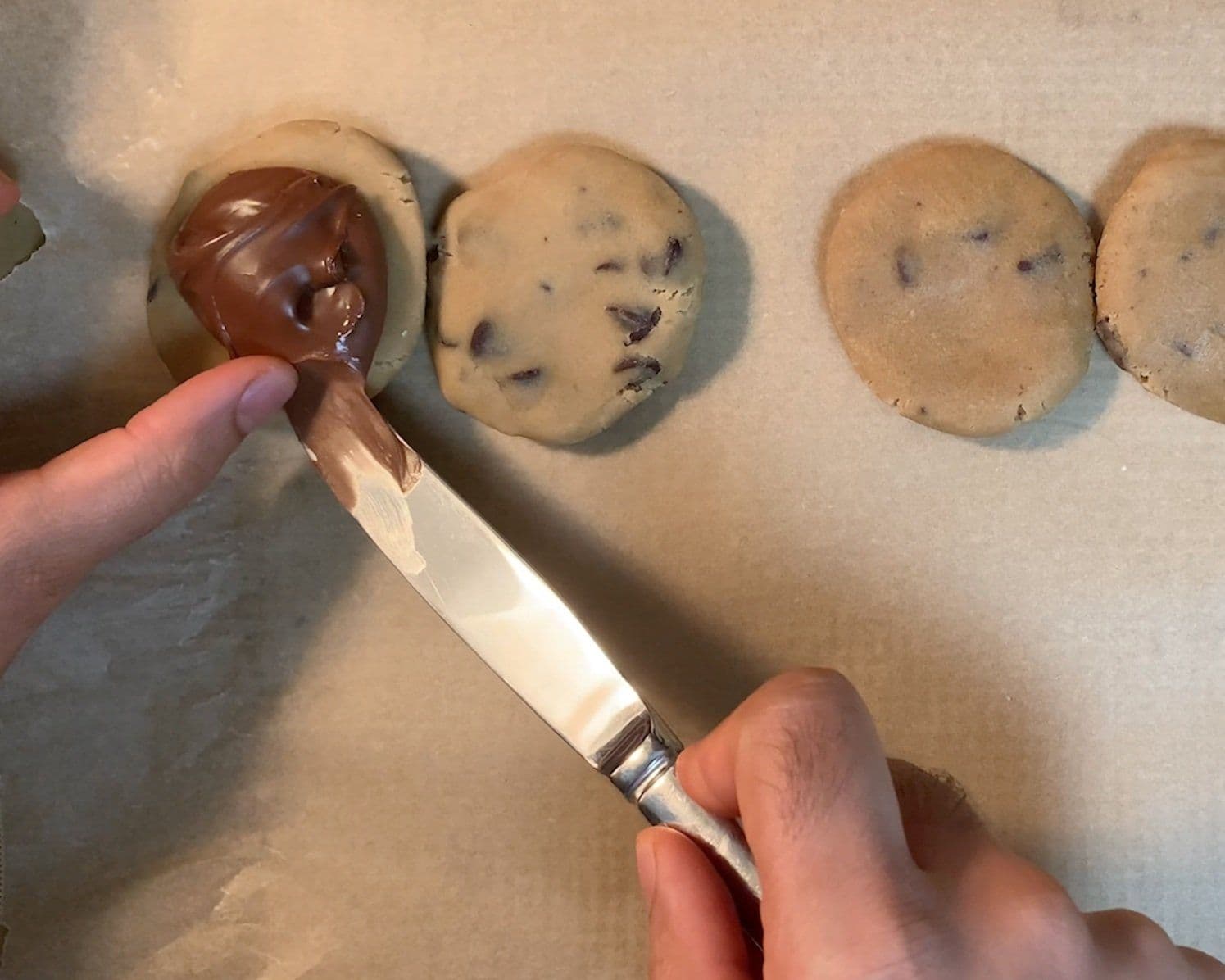 Hand holding a brush applying chocolate coating to a stuffed cookie, with additional plain cookies on parchment paper.