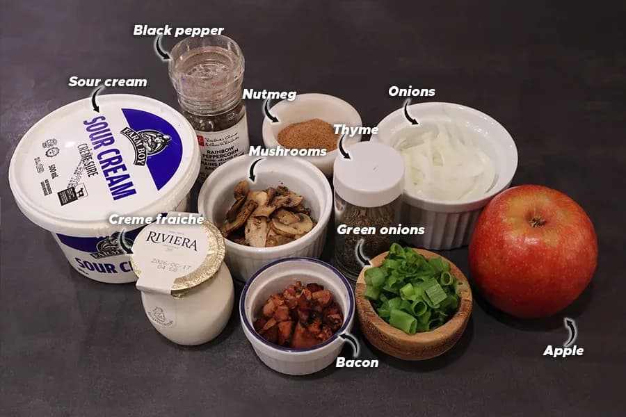 Ingredients for Flammkuchen toppings displayed on dark surface: sour cream, crème fraîche, mushrooms, onions, thyme, nutmeg, black pepper, green onions, bacon, and apple.