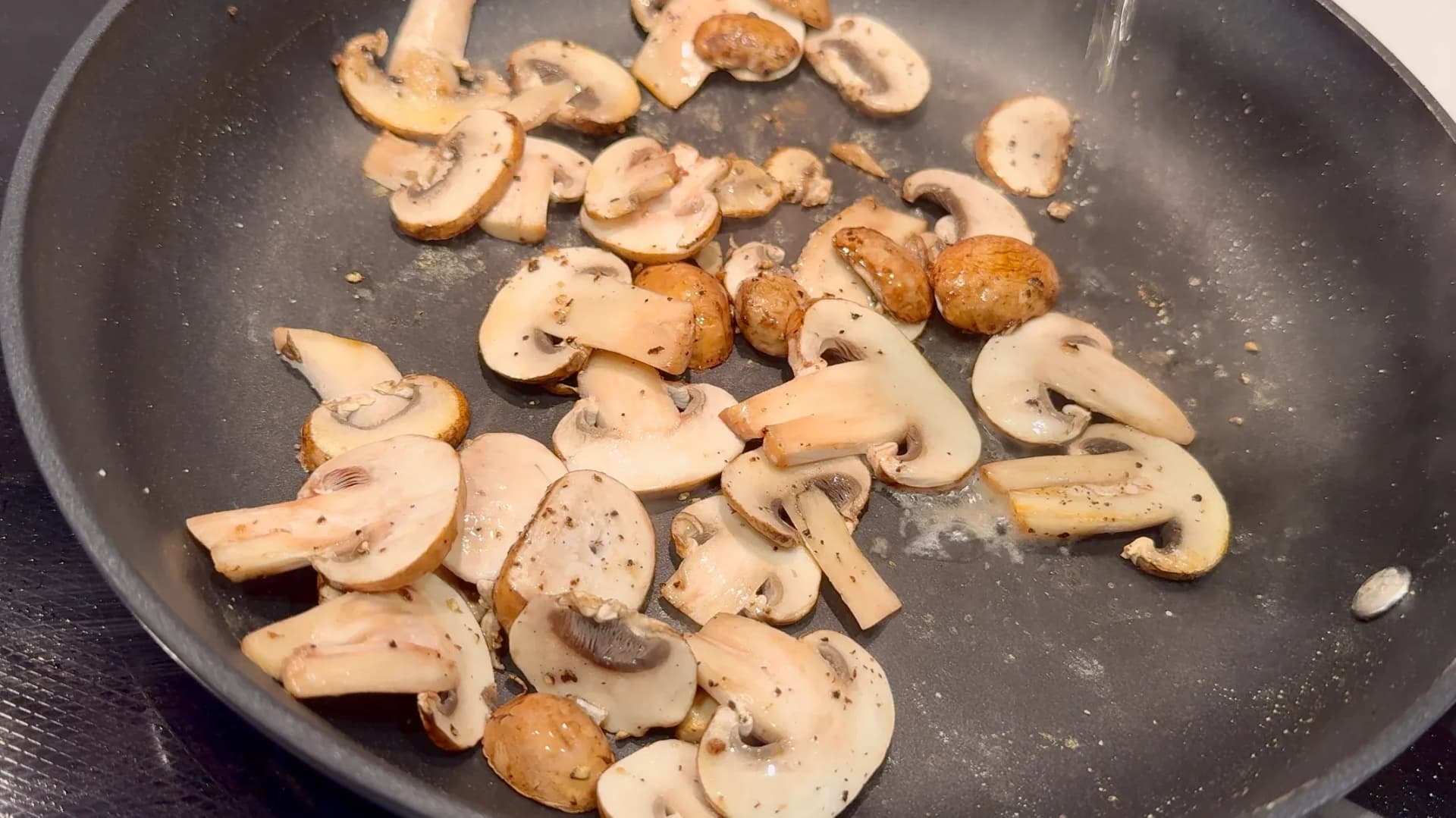 Sliced mushrooms sautéing in a dark pan with butter and seasonings for vegetarian Flammkuchen topping.