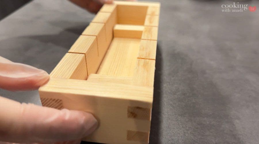 Wooden oshizushi mold lined with plastic wrap, showing compartments for layering pressed sushi ingredients.