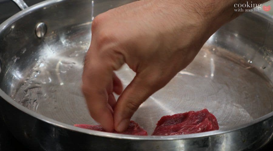 Hand searing a red beef cube in a hot stainless steel pan for Balinese beef rendang.