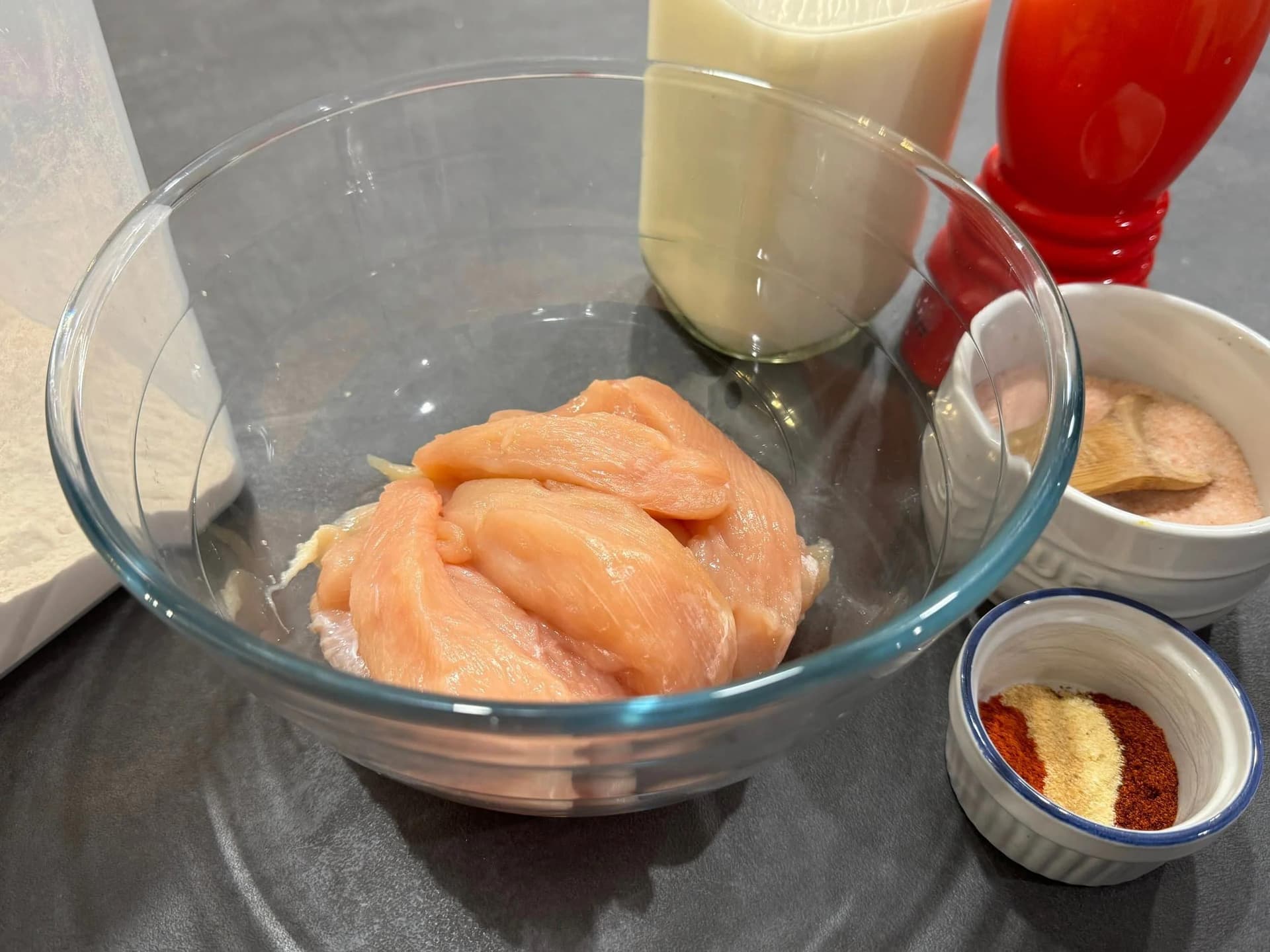 Raw chicken breast strips marinating in buttermilk with spices in a glass bowl, surrounded by ingredient bowls.