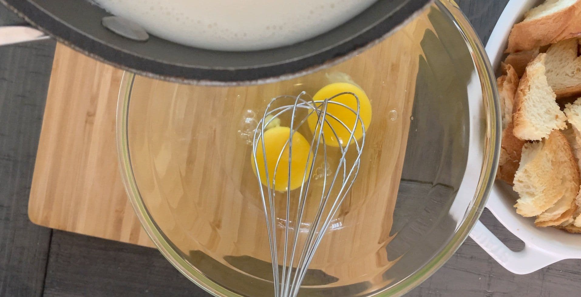 Whisking egg yolk in a glass bowl with a wire whisk, brioche bread cubes visible on the side.