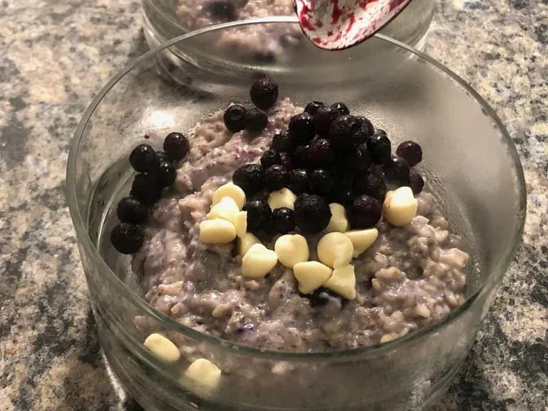 Purple oatmeal in a glass bowl topped with blueberries and white chocolate chips on granite countertop.