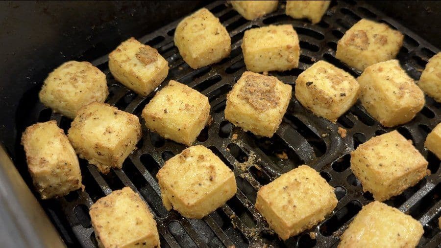 Golden-brown tofu cubes crisping in an air fryer basket halfway through cooking.