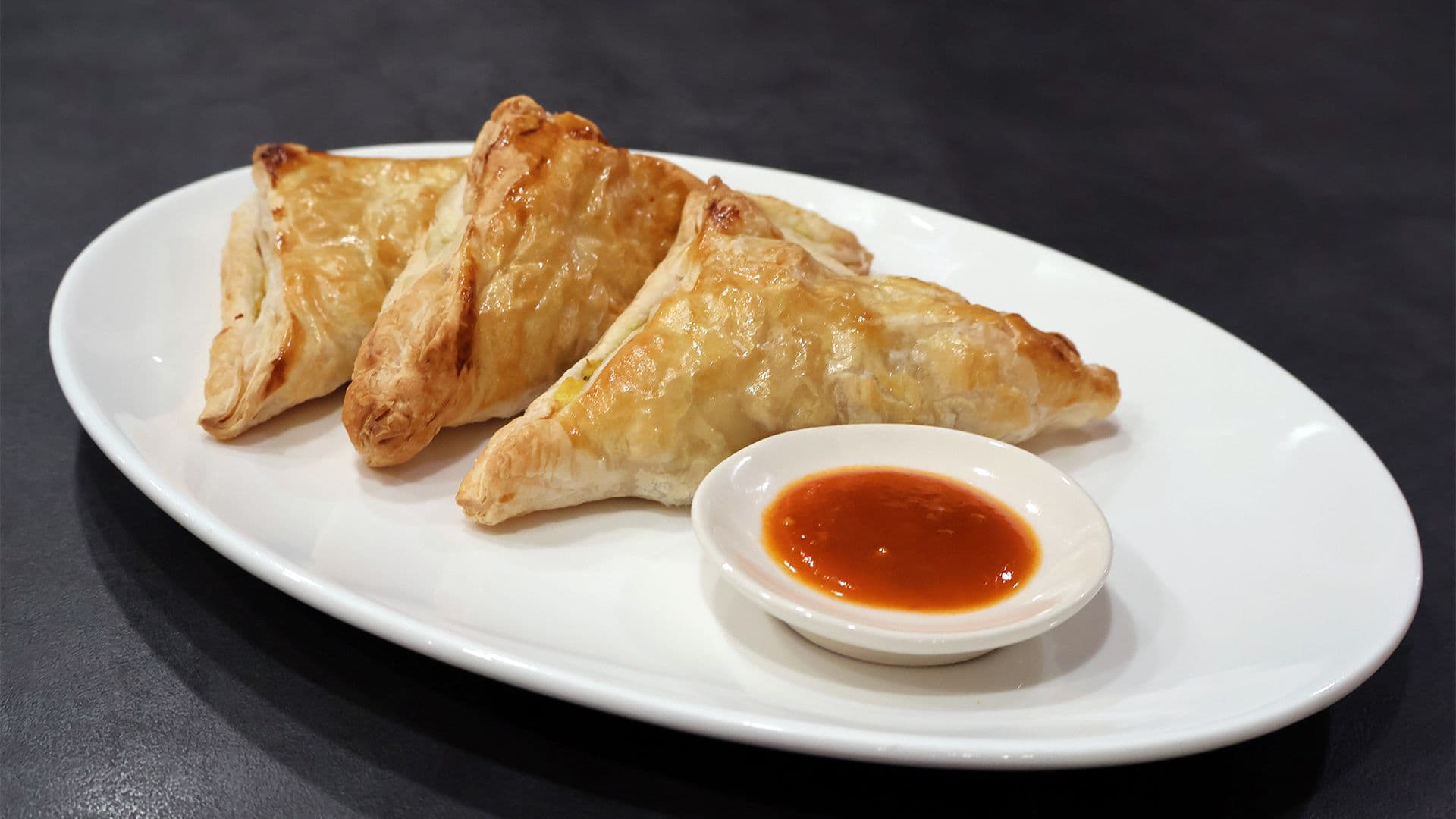 Three golden-brown aloo patties on a white plate with a small bowl of chutney for dipping.
