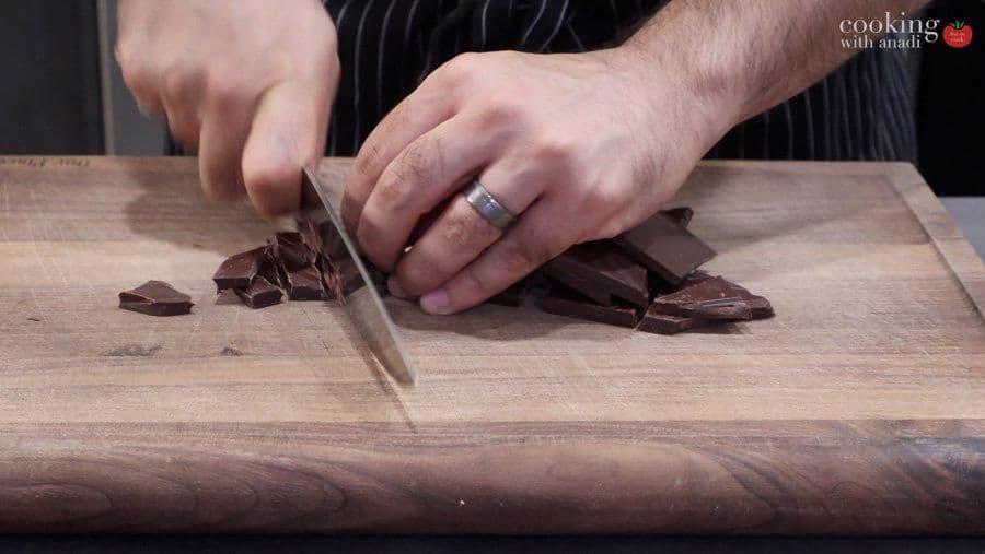 Chopping dark chocolate into small pieces on a wooden cutting board with a chef's knife.