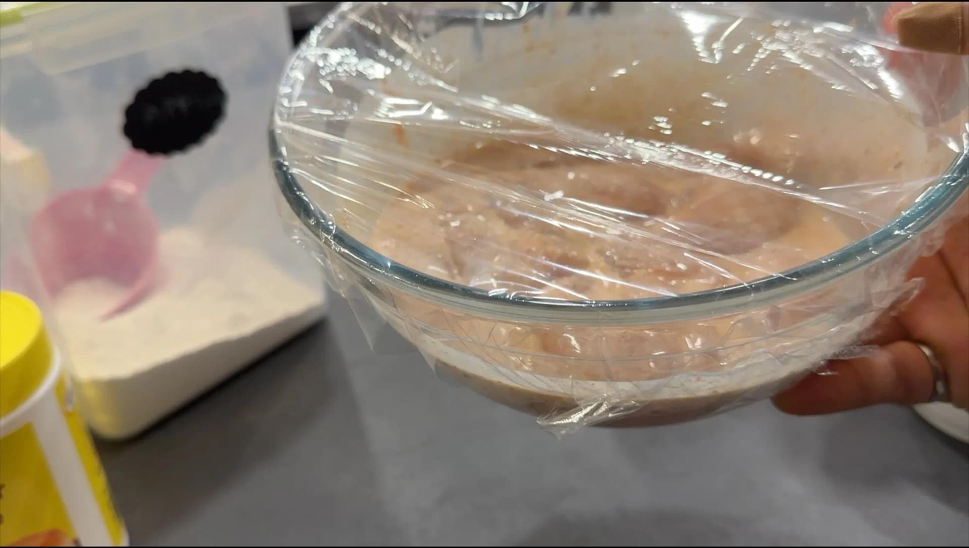 Chicken pieces marinating in buttermilk in a glass bowl covered with plastic wrap.