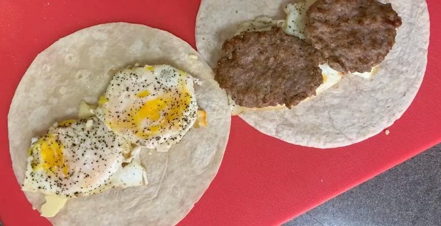 Two assembled breakfast wraps on a red surface with eggs and beef patties visible inside flour tortillas.