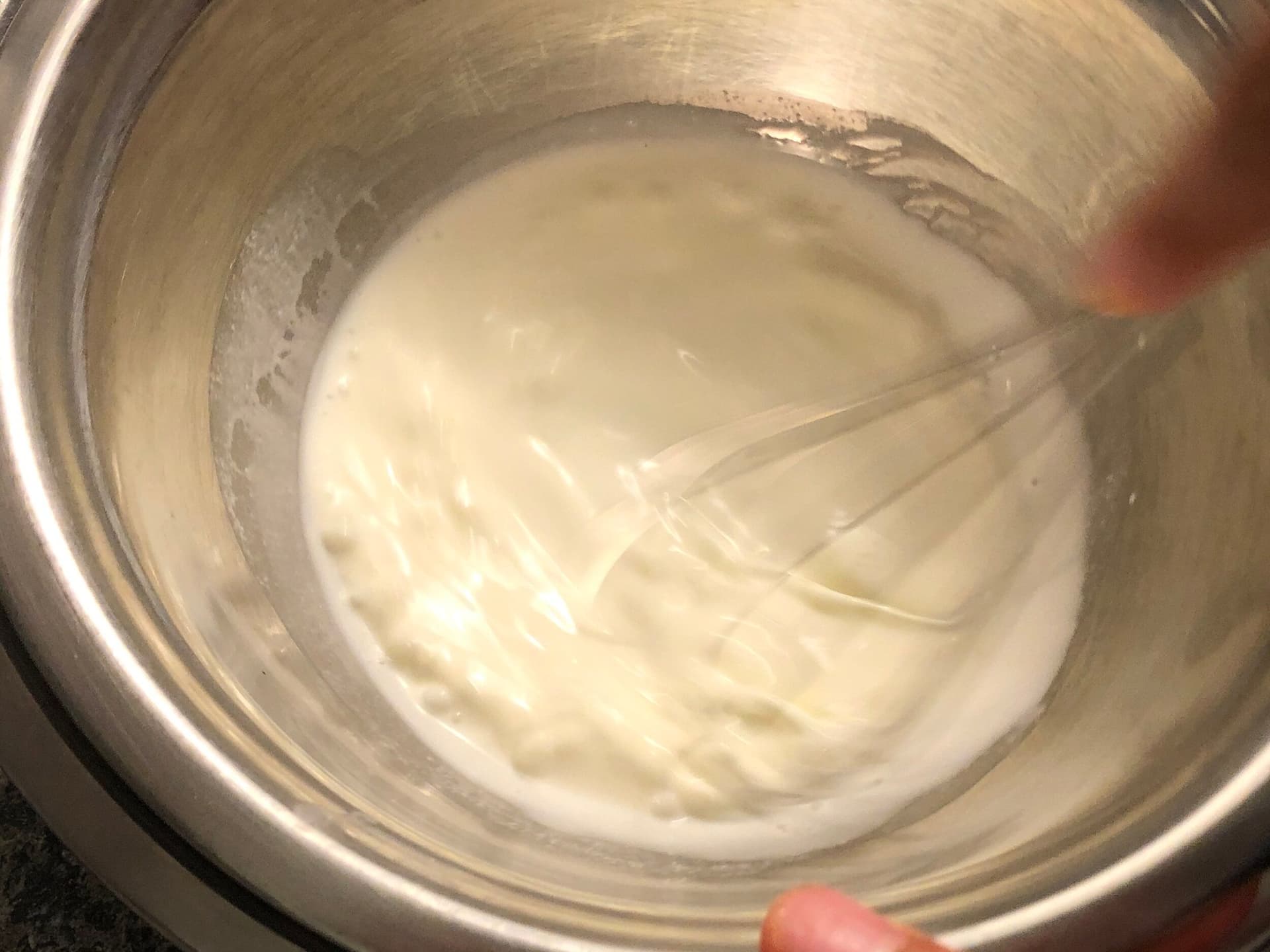 Whipped cream being whisked by hand in a metal bowl, showing soft peaks forming on the whisk.