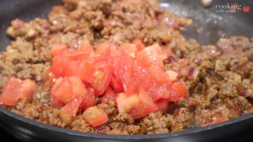 Ground beef cooked with diced tomatoes in a dark pan, showing the tomato pieces being incorporated into the meat mixture.