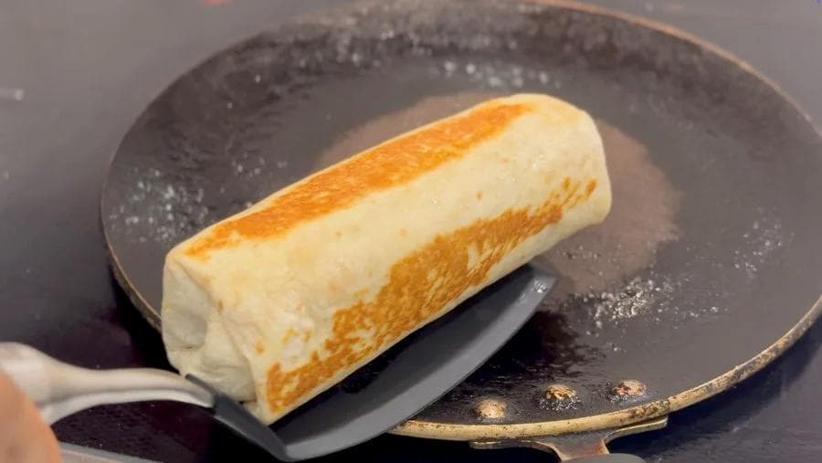 Toasted breakfast burrito with golden brown exterior in a dark skillet with a spatula.