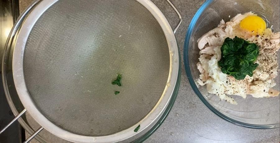 Drained spinach in a fine mesh strainer next to a bowl with ricotta, egg, chicken, and fresh spinach ready to mix.