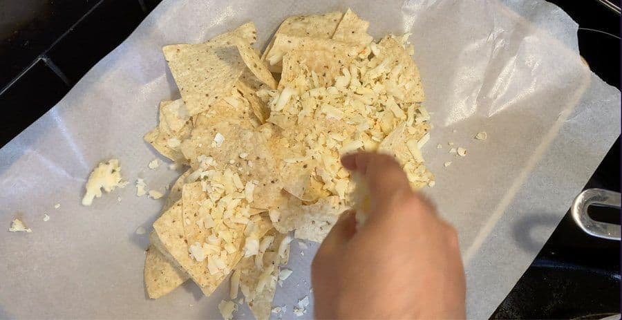 Hand sprinkling shredded cheese over tortilla chips on a parchment-lined baking sheet.