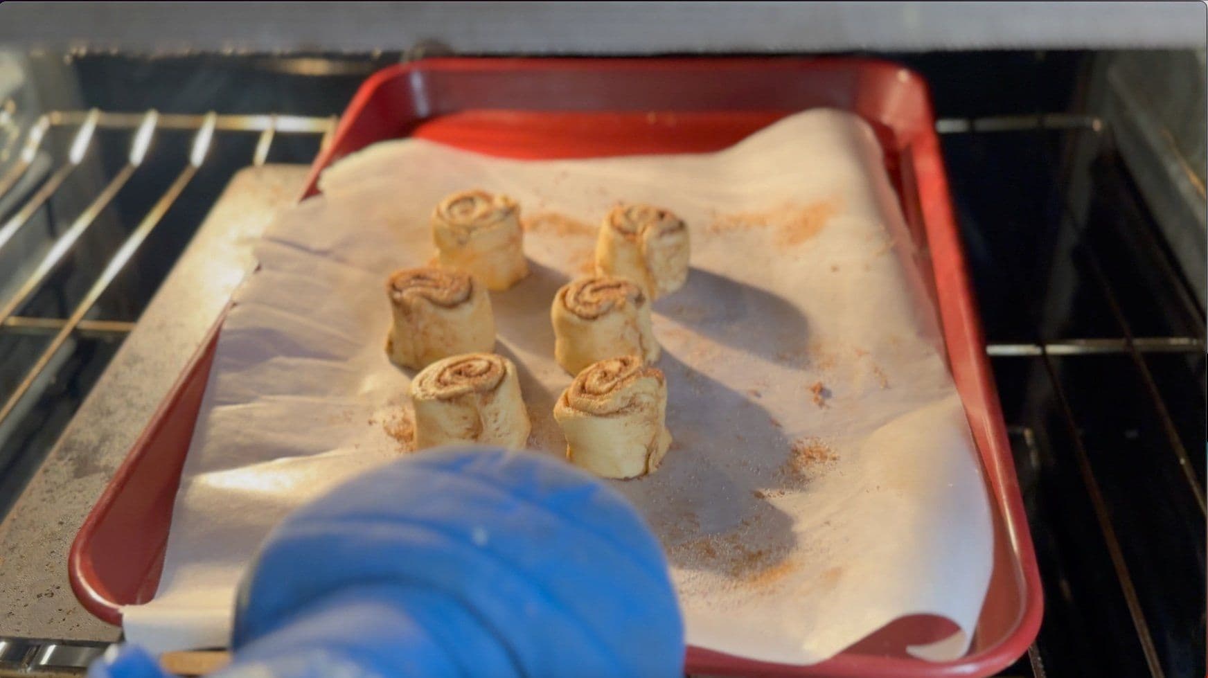 Six golden-brown cinnamon roll pinwheels on parchment paper in a red baking tray in the oven.