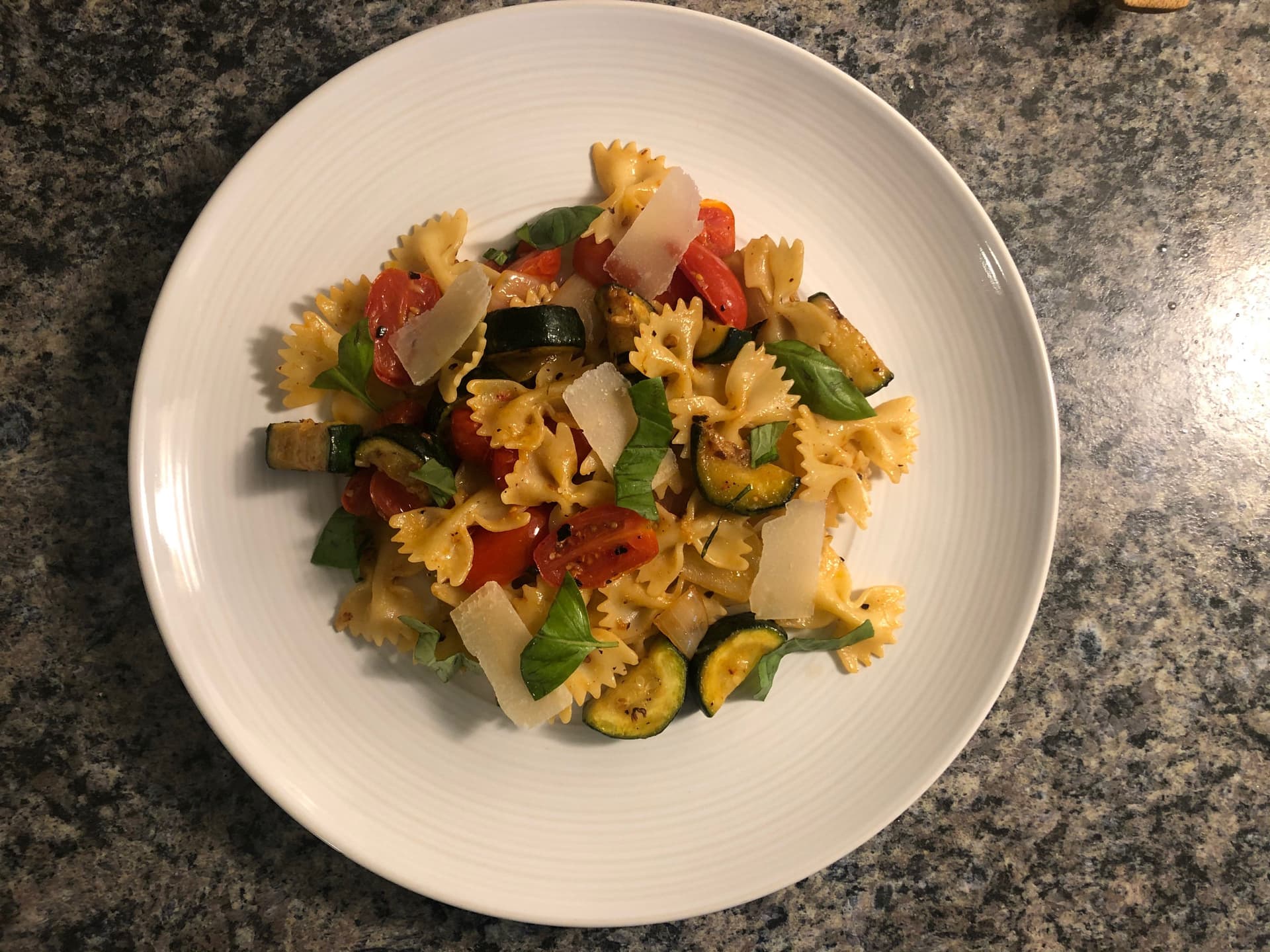 Mediterranean pasta salad with bow-tie pasta, grilled zucchini, red peppers, mozzarella, and fresh basil on white plate.