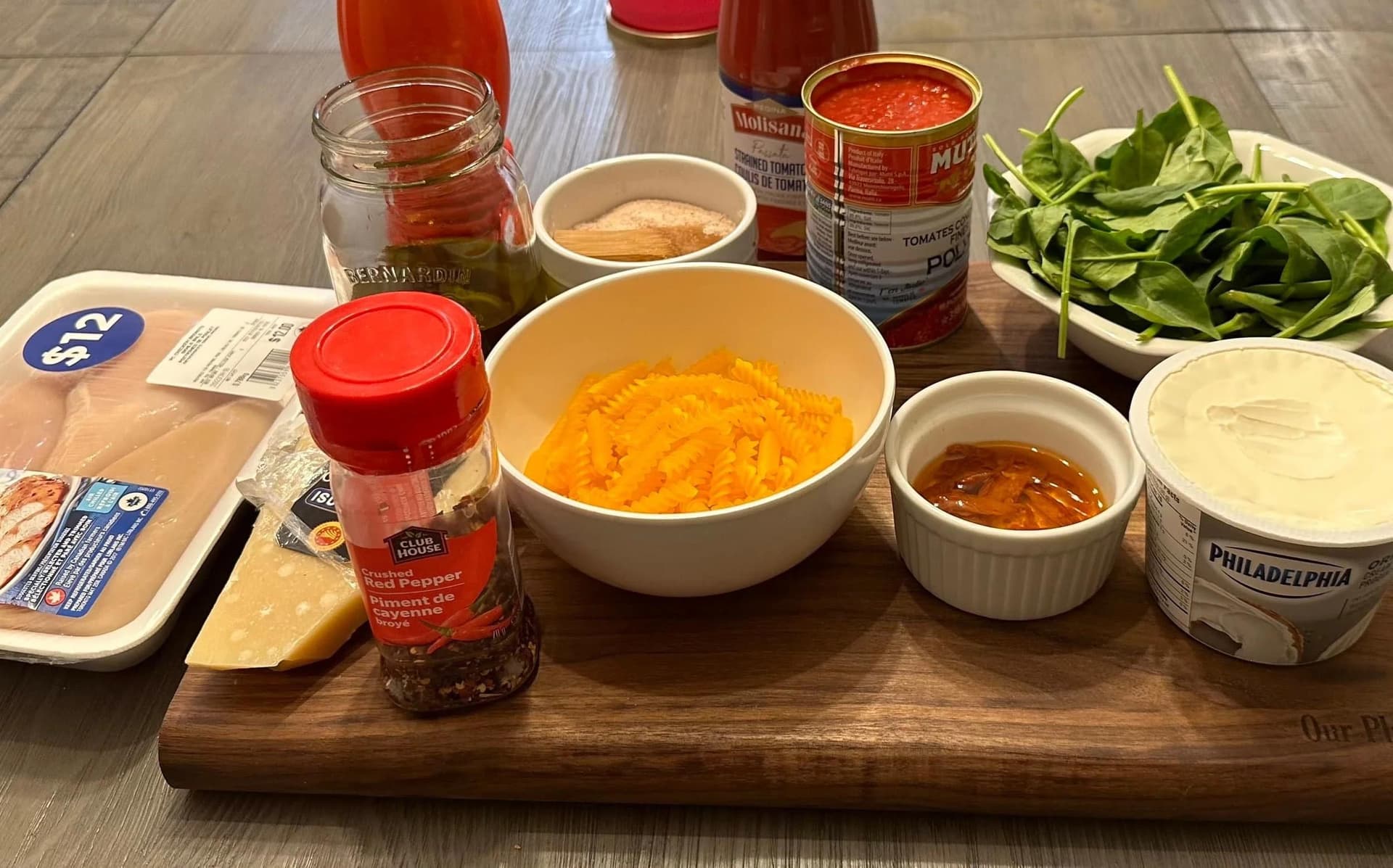 Ingredients for creamy tomato chicken pasta laid out on a wooden board, including shredded cheese, tomato sauce, cream cheese, and fresh spinach.