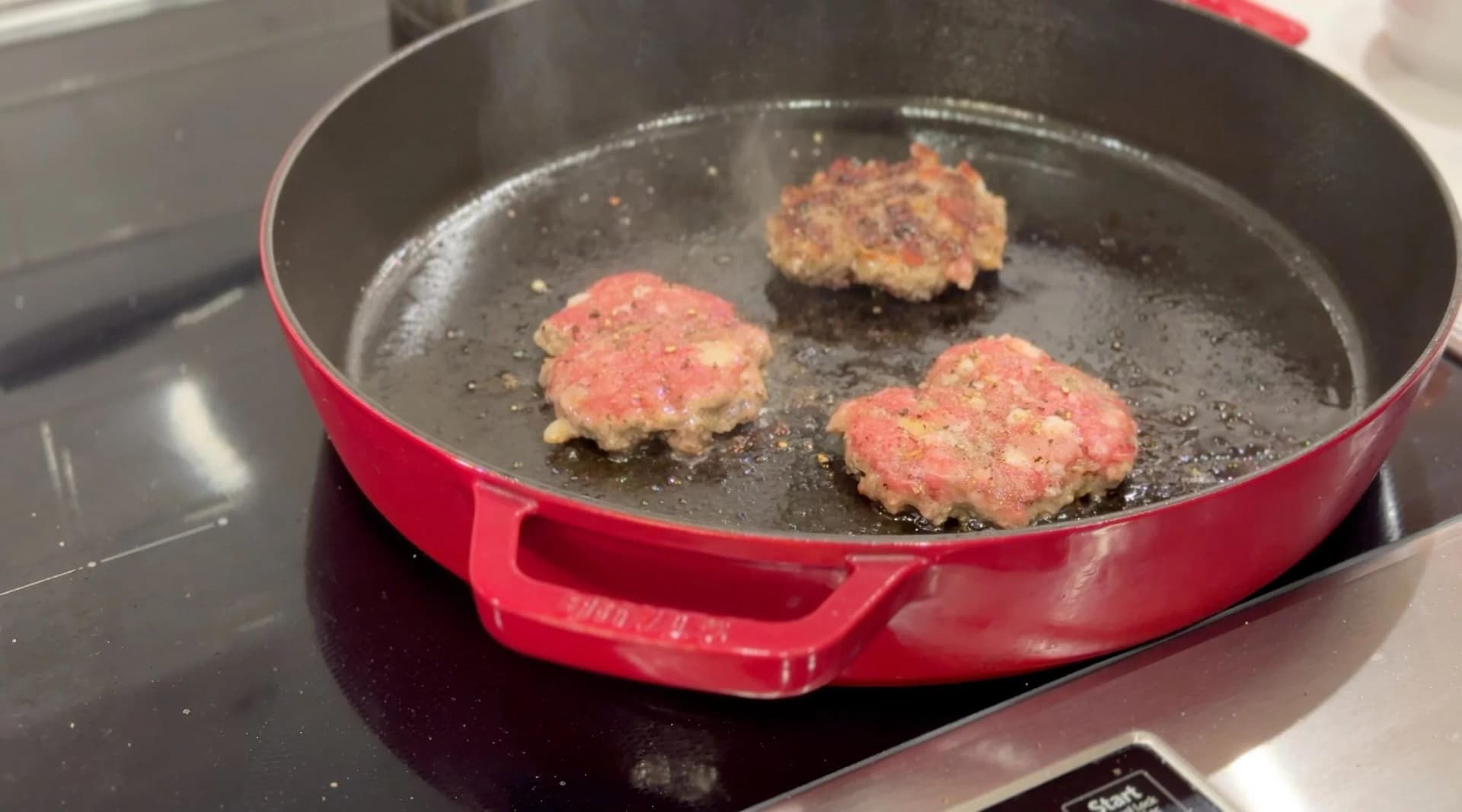 Four raw burger patties cooking in a red cast iron pan on a stovetop with oil.