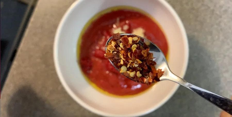 Red pizza sauce in white bowl with chili oil drizzle and red pepper flakes on spoon.
