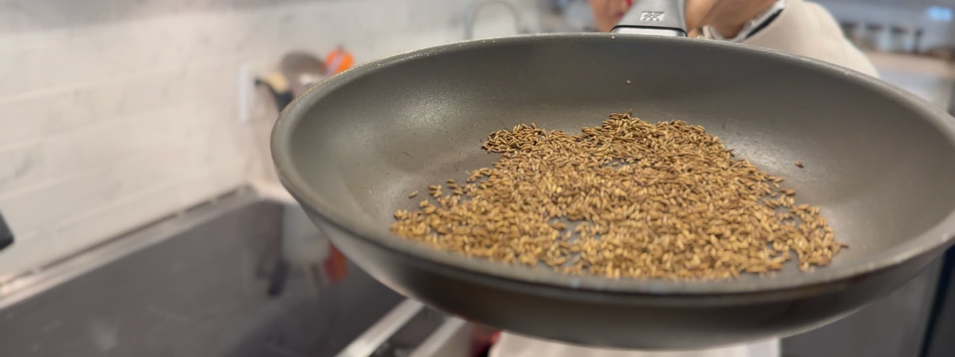 Cumin seeds being roasted in a gray pan on low heat, turning golden brown in color.