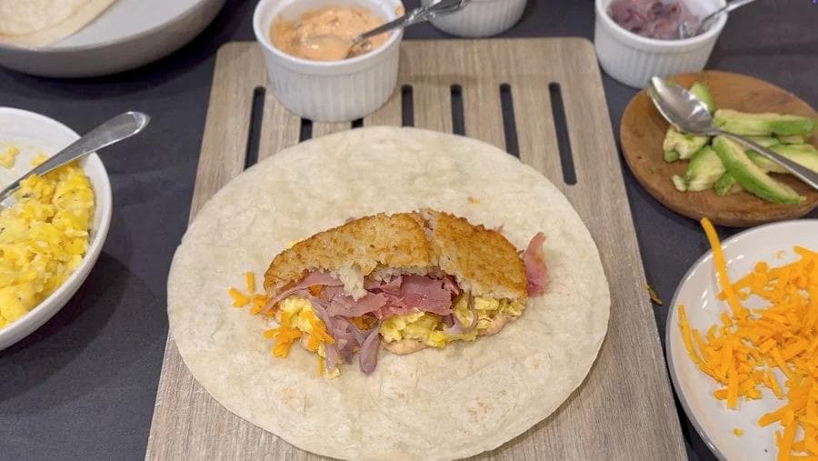 Assembling a breakfast burrito with hash browns, scrambled eggs, and ham on a flour tortilla surrounded by ingredient bowls.