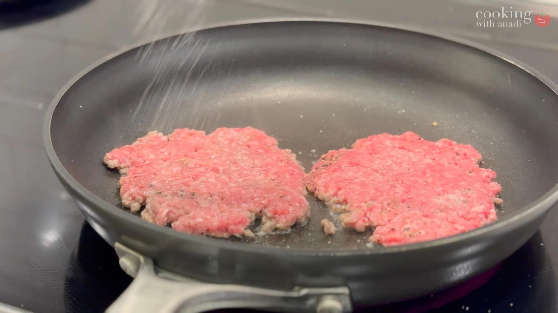 Two pink ground beef patties being cooked in a nonstick skillet with onions on top.