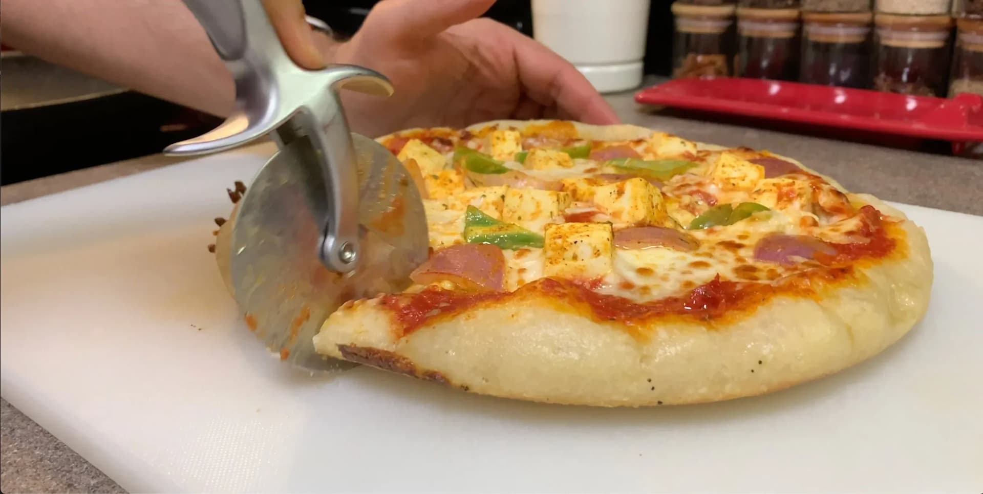 Paneer tikka pan pizza being cut with a server, showing melted cheese and toppings on a white plate.