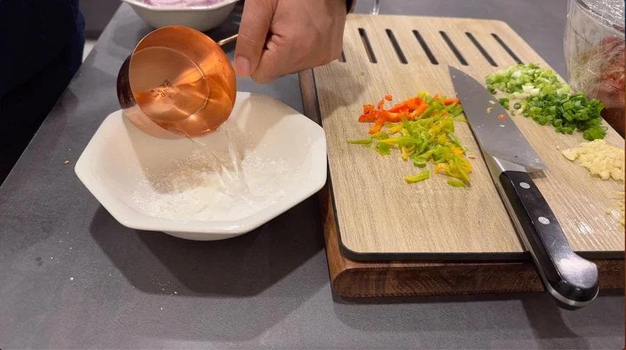 Pouring cornstarch slurry into a white bowl while chopped vegetables sit on cutting boards nearby.