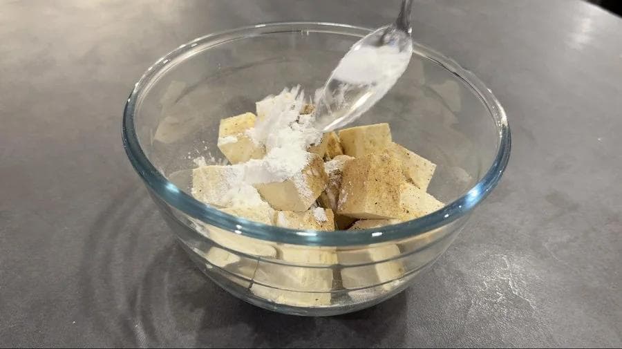 Tofu cubes coated with white cornstarch and seasonings in a glass bowl with a spoon.