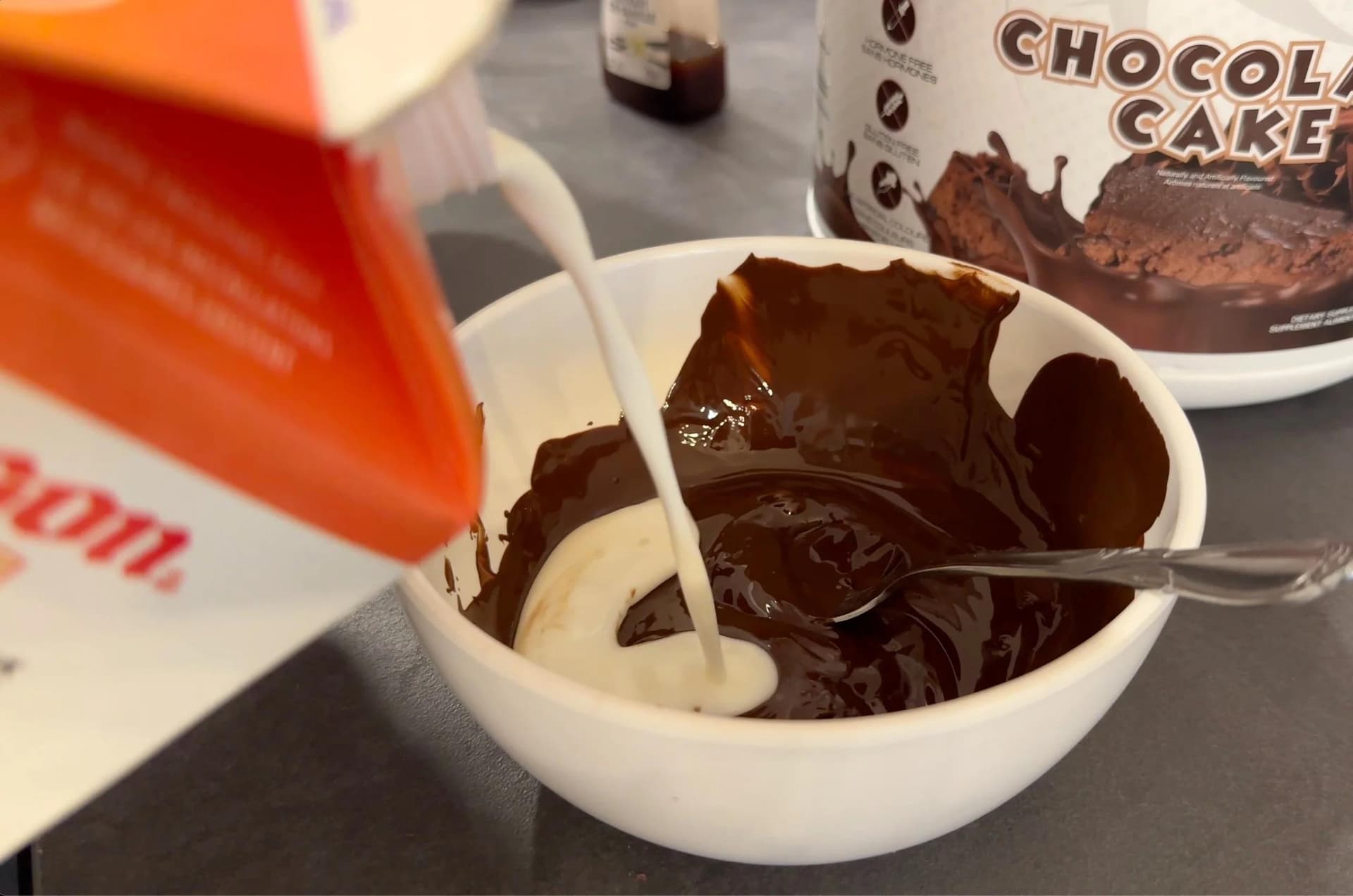 Milk being poured into melted chocolate and protein powder in a white bowl with a spoon.