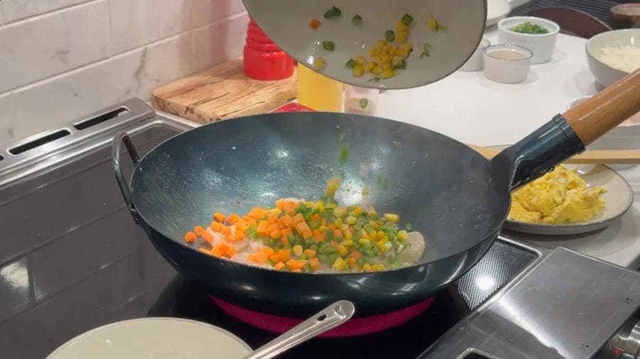 Diced carrots and peas stir-frying in a wok with oil, mid-cooking stage of fried rice preparation.