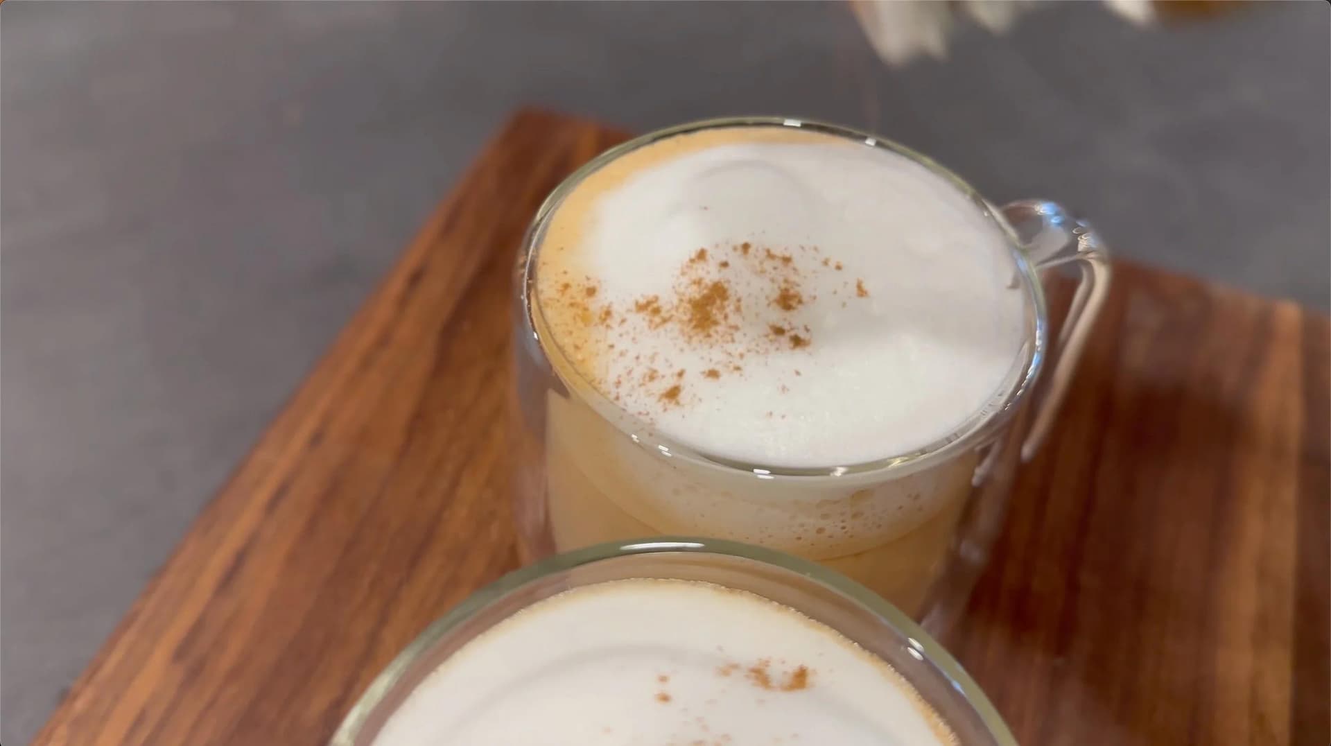 Two mugs of dirty chai latte topped with frothed milk and cinnamon powder on a wooden board.