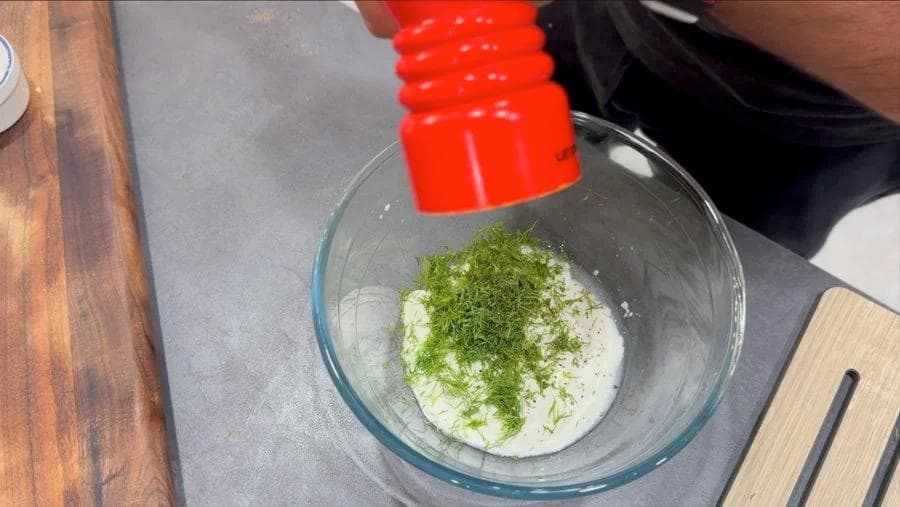Fresh dill sprinkled over creamy yogurt and vinegar mixture in a glass bowl for cucumber salad dressing.