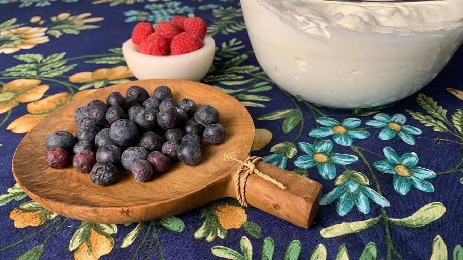 Homemade whipped cream in a glass bowl with fresh blueberries on a wooden plate and raspberries in a small white bowl.