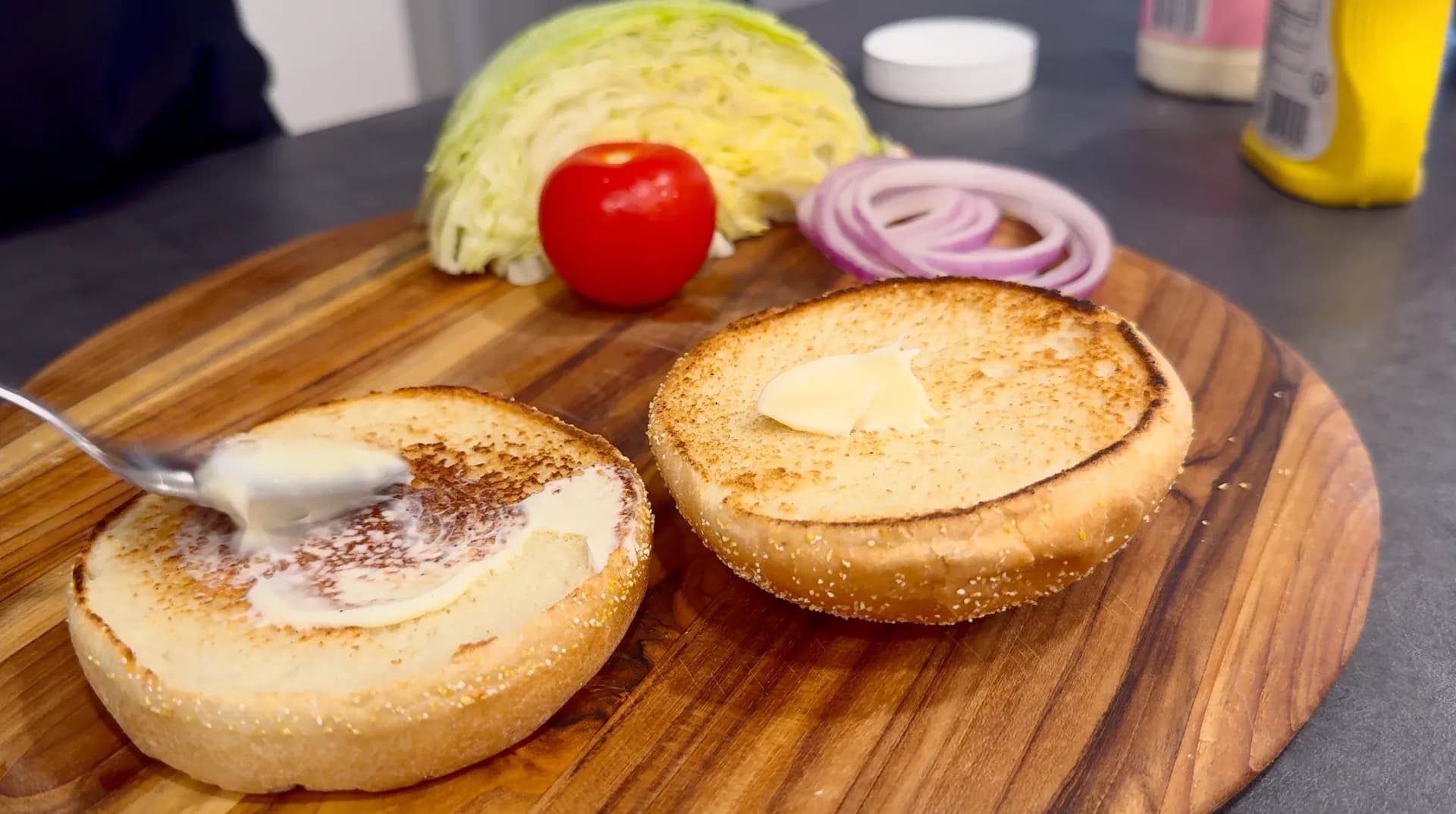 Toasted burger buns spread with mayonnaise on a wooden board with fresh tomato, cabbage, and red onion slices nearby.