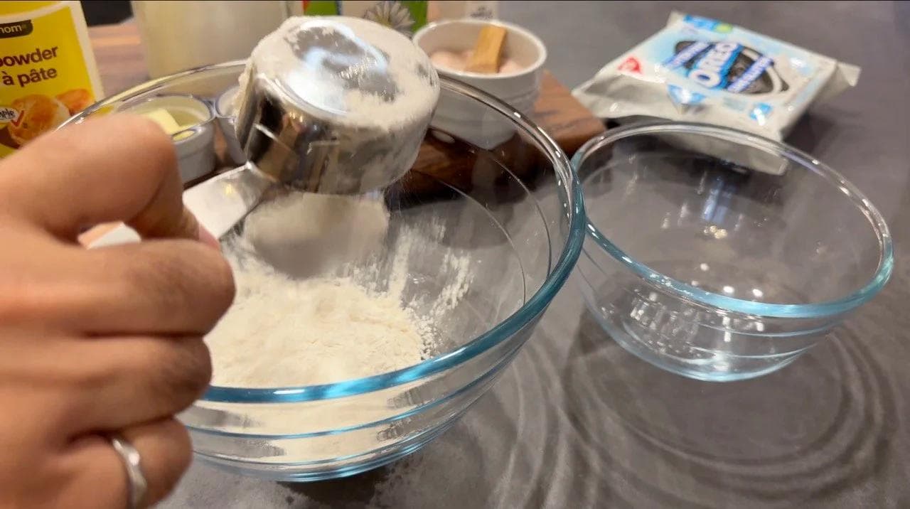 Dry ingredients being scooped into a glass mixing bowl during pancake batter preparation.