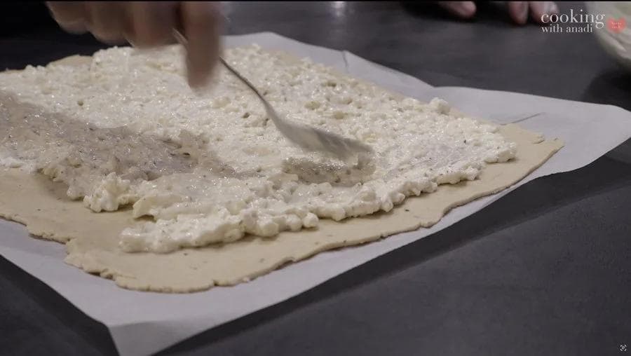 Spreading crumbly cheese filling evenly over thin dough with a spatula, leaving borders.